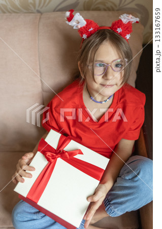 A girl in a Santa Claus hat opens a gift. It's Christmas time. A girl in a Santa Claus hat opens a gift. It's Christmas time. 133167659