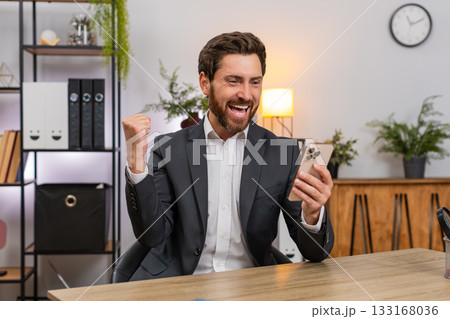 Middle-aged businessman texting happy news on smartphone celebrates project win with victory gesture Middle-aged businessman texting happy news on smartphone celebrates project win with victory gesture 133168036