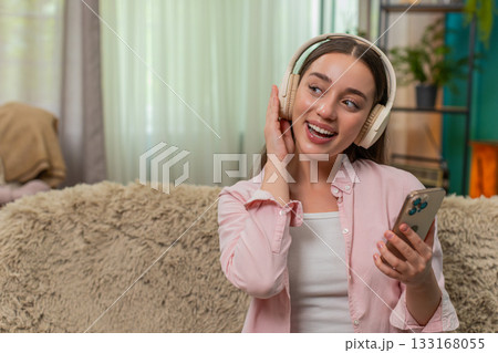 Young woman dancing on sofa wearing wireless headphones enjoying favorite music with joyful energy Young woman dancing on sofa wearing wireless headphones enjoying favorite music with joyful energy 133168055