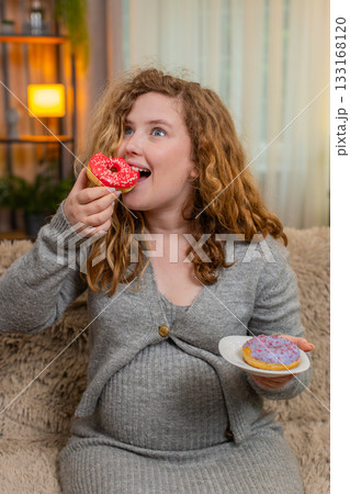 Pregnant young woman eating donuts on home sofa laughing with joyful expression enjoying sweet treat 133168120