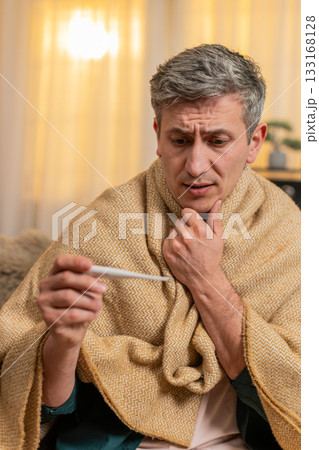 Young man checks body temperature with thermometer looks worried about possible illness and fever Young man checks body temperature with thermometer looks worried about possible illness and fever 133168128