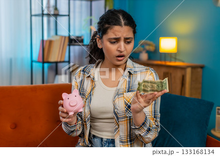 Woman sadly looking at piggy bank with one dollar inside financial difficulties limited savings Woman sadly looking at piggy bank with one dollar inside financial difficulties limited savings 133168134