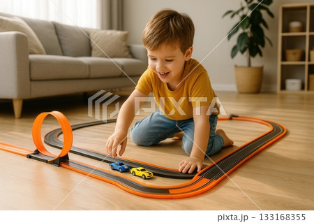 A cheerful child enjoys racing toy cars on an orange track with loops in a bright, modern living room. Great for themes of play, creativity, childhood fun, and toy advertising. 133168355