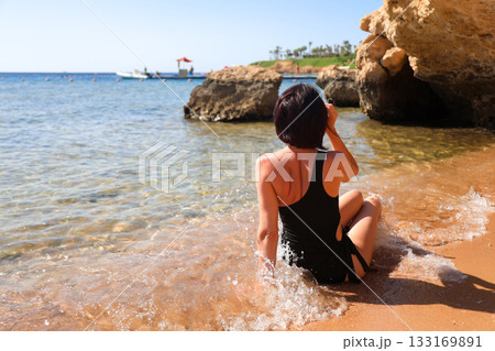 A brunette woman with glasses in a black bathing suit sits on the sand overlooking the sea, view from the back A brunette woman with glasses in a black bathing suit sits on the sand overlooking the sea, view from the back 133169891