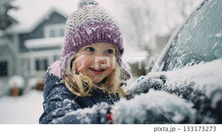 A cheerful toddler in a knitted hat brushes snow from a car on a winter day 133173273