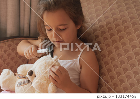Playing doctor: a small girl examines a teddy bear's ear with an otoscope Playing doctor: a small girl examines a teddy bear's ear with an otoscope 133174458