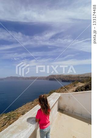 Child enjoying the stunning cliffside view over the water on a sunny day in the picturesque Greek island of Santorini 133174488
