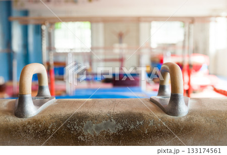 Pommel horse in gymnasium. The horse is tan leather with wooden legs and handles. 133174561