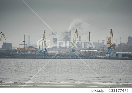 Industrial landscape, a factory with pipes and cranes in the port against a gray foggy sky Industrial landscape, a factory with pipes and cranes in the port against a gray foggy sky 133177473