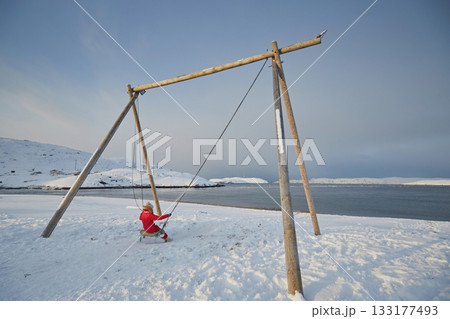 A girl in a red jumpsuit swings on a large swing against the backdrop of the non-freezing northern sea and snow in winter 133177493