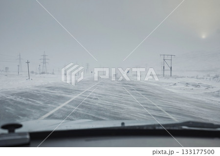 Sweeping snow on the road in a stormy wind. View from the windshield of a car on a snowdrift against a sky with fog. Dangerous driving in winter in snowfall 133177500