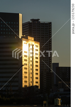 多摩川の向かい側の川崎のビル群の間からさす快晴の空に沈みゆく夕方の太陽と多摩川の河川敷 多摩川の向かい側の川崎のビル群の間からさす快晴の空に沈みゆく夕方の太陽と多摩川の河川敷 133179230