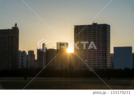 多摩川の向かい側の川崎のビル群の間からさす快晴の空に沈みゆく夕方の太陽と多摩川の河川敷 133179231