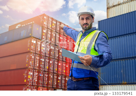 Portrait of handsome caucasian man with white helmet and reflection shirt, holding document paper and pointing finger in front of container cargo store. import and export logistic industry concept. 133179541