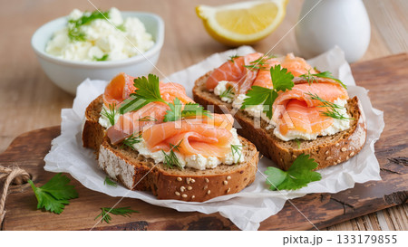 Open sandwiches with smoked salmon, cottage cheese, dill and parsley on whole grain bread, served on wooden board with parchment. Fresh and healthy snack. Open sandwiches with smoked salmon, cottage cheese, dill and parsley on whole grain bread, served on wooden board with parchment. Fresh and healthy snack. 133179855