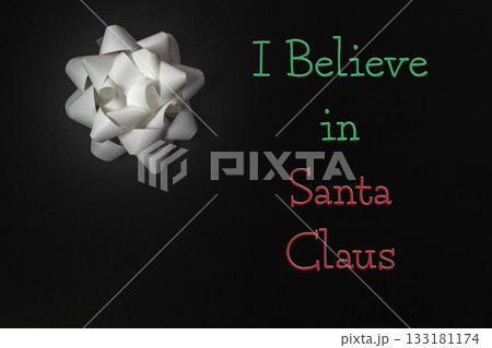White gift bow on dark background with festive text expressing belief in Santa Claus 133181174