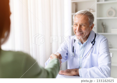 Smiling senior male doctor shaking hands with female patient in bright clinic 133182025
