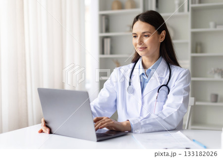 Smiling female doctor in white coat working on laptop in clinic office Smiling female doctor in white coat working on laptop in clinic office 133182026
