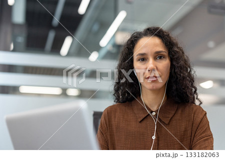 Woman with curly hair in brown shirt wearing earphones, looking at camera while using laptop for a focused online video conference in a modern office setting Woman with curly hair in brown shirt wearing earphones, looking at camera while using laptop for a focused online video conference in a modern office setting 133182063