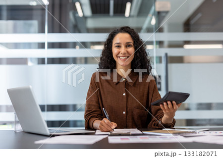 Young entrepreneur working on financial calculations and paperwork at office desk, holding a calculator and writing notes, exuding confidence and professionalism Young entrepreneur working on financial calculations and paperwork at office desk, holding a calculator and writing notes, exuding confidence and professionalism 133182071