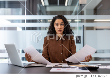 Stressed businesswoman sitting at a modern office desk, looking directly at the viewer with a concerned expression while holding papers, signifying workload, financial problems, and confusion 133182072
