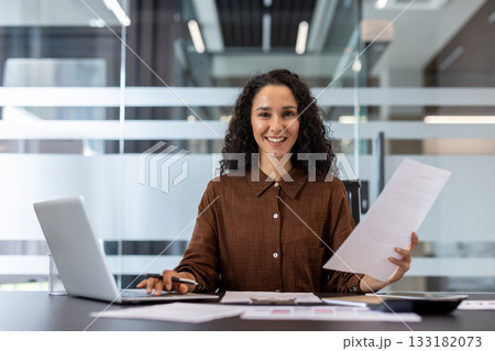 Female professional sitting at office desk, holding documents, and using a laptop, representing modern business, career success, and productivity in a corporate environment Female professional sitting at office desk, holding documents, and using a laptop, representing modern business, career success, and productivity in a corporate environment 133182073