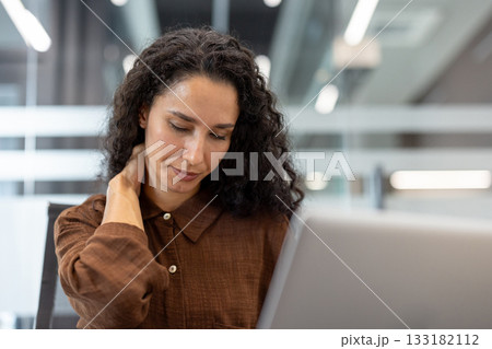 Young woman feeling intense neck pain and tension while working on her laptop in a modern office, symbolizing stress, exhaustion, and health issues from an overworked lifestyle 133182112