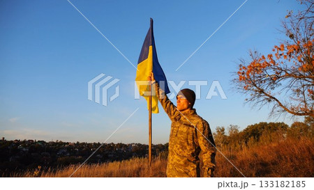 Male military in uniform lifting blue-yellow banner in honor of the victory against russian aggression at sunset. Young soldier of ukrainian army raised a waving flag of Ukraine at countryside Male military in uniform lifting blue-yellow banner in honor of the victory against russian aggression at sunset. Young soldier of ukrainian army raised a waving flag of Ukraine at countryside 133182185