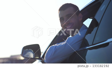 Young smiling businessman in shirt leaning out of driver window at modern electric car and looking into camera. Portrait of handsome successful entrepreneur in innovative electrical vehicle. Slow mo 133182192