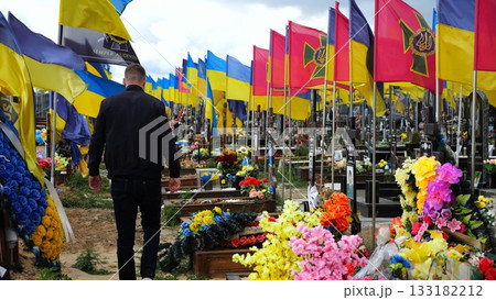Mourning man go among countless graves of ukrainian soldiers with national flags at military cemetery in Kharkiv. Scene reflects personal sorrow and nation collective loss caused by russian aggression Mourning man go among countless graves of ukrainian soldiers with national flags at military cemetery in Kharkiv. Scene reflects personal sorrow and nation collective loss caused by russian aggression 133182212