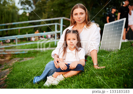 Mother and daughter sit together on grass, embracing in warm moment with solar panel beside in the evening. Concept of renewable energy and family, highlighting commitment to sustainable future. Mother and daughter sit together on grass, embracing in warm moment with solar panel beside in the evening. Concept of renewable energy and family, highlighting commitment to sustainable future. 133183183