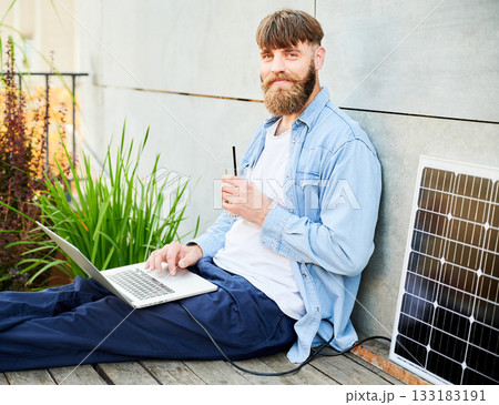 Portrait of barded man works on laptop on lap with glass of iced coffee. Solar panel nearby, emphasizing sustainable, modern eco-friendly workspace that combines technology and sustainability. Portrait of barded man works on laptop on lap with glass of iced coffee. Solar panel nearby, emphasizing sustainable, modern eco-friendly workspace that combines technology and sustainability. 133183191