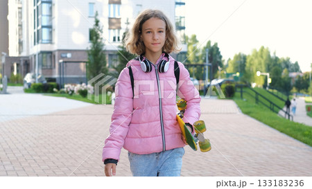 Teen girl walks to school in the morning carrying a colorful skateboard wearing a pink jacket and backpack with headphones around her neck 133183236
