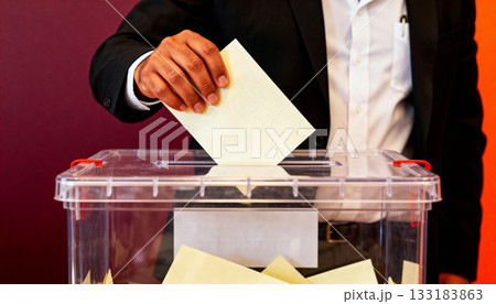 man's hand dropping a ballot into a transparent ballot box during an election, symbolizing the importance of democratic procedures and local government elections 133183863