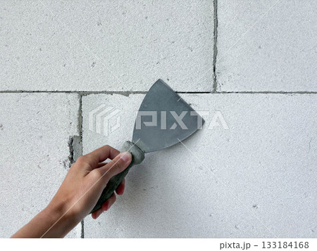 A hand with a spatula on a white block wall. Preparing for interior wall finishing. Renovation and construction. A hand with a spatula on a white block wall. Preparing for interior wall finishing. Renovation and construction. 133184168