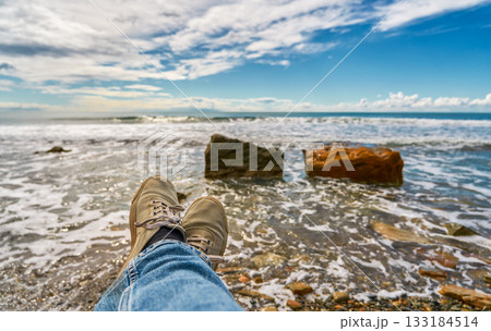 Feet in sneakers against the backdrop of the surf and a rocky seashore. A magnificent view and space for rest and relaxation after the work week, weekend by the sea concept 133184514