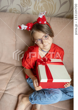 A girl in a Santa Claus hat opens a gift. It's Christmas time. A girl in a Santa Claus hat opens a gift. It's Christmas time. 133184700