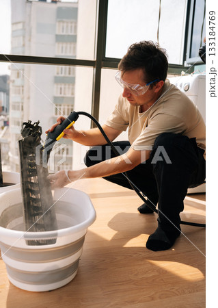 Vertical shot of professional cleaning service worker wearing safety glasses using steam cleaner to clean air conditioner filter in bucket of water, performing maintenance on HVAC system. Vertical shot of professional cleaning service worker wearing safety glasses using steam cleaner to clean air conditioner filter in bucket of water, performing maintenance on HVAC system. 133184769