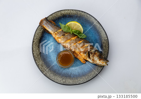 Whole grilled sea bass with lemon, basil and sauce on a blue ceramic plate, top view Whole grilled sea bass with lemon, basil and sauce on a blue ceramic plate, top view 133185500