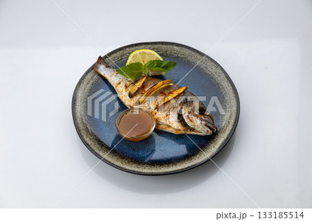 Whole grilled dorado with lemon, basil and sauce on a blue ceramic plate, white background 133185514