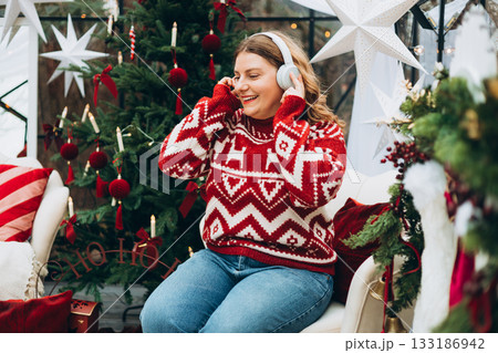Cute young smiling woman listening music from smartphone during cozy Xmas holidays at home. Smiling Caucasian millennial lady in headphones sits near a Christmas tree Cute young smiling woman listening music from smartphone during cozy Xmas holidays at home. Smiling Caucasian millennial lady in headphones sits near a Christmas tree 133186942