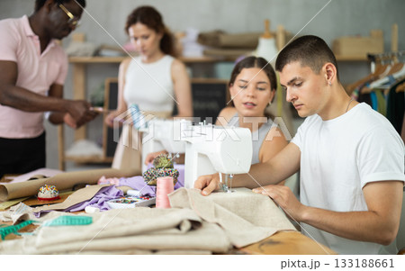 Young man sewing while others working with sewing-machine and making details Young man sewing while others working with sewing-machine and making details 133188661