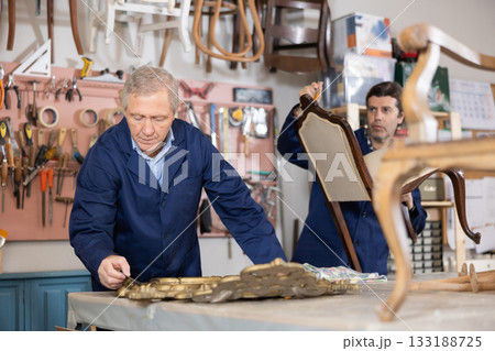 Mature male carpenter and his apprentice working on restoring vintage mirror and chair in workshop Mature male carpenter and his apprentice working on restoring vintage mirror and chair in workshop 133188725