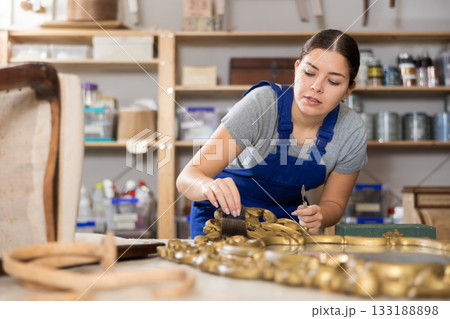 Girl restorer carefully covers carved wooden frame of antique wall mirror with thin layer of varnish 133188898