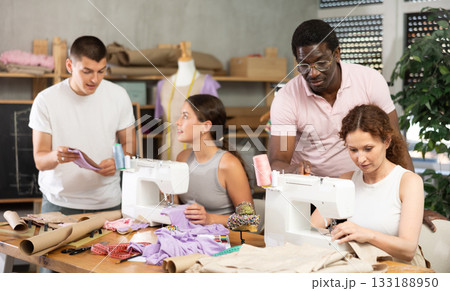 During lesson, African man teacher helps student sew, others work with fabric, blank, dress patterns 133188950