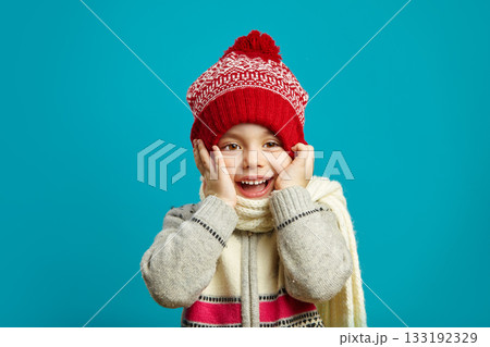 Funny girl in winter hat, put her hands to cheeks, cute smile, expresses a cheerful mood and surprise, children's emotional portrait on blue background. 133192329
