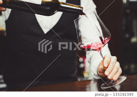 Close-up photo, sommelier pours red wine to glass on bar counter background. 133192395