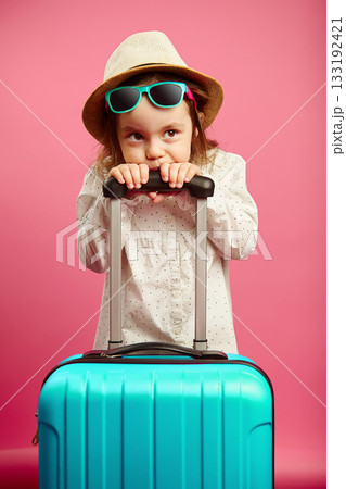 Bored little girl with blue suitcase, wears a straw hat and sunglasses, expresses puzzlement and confusion, portrait of child on isolated pink isolated. 133192421