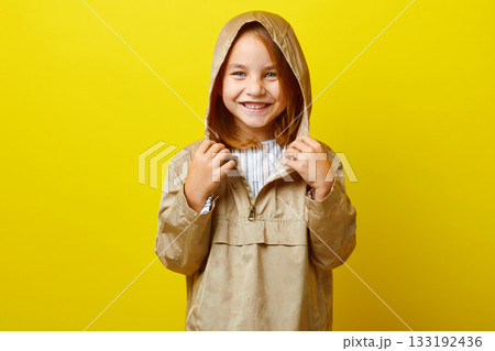 Cheerful portrait of happy little girl in demi-season beige jacket with hood from rain, studio shot on colored background. 133192436