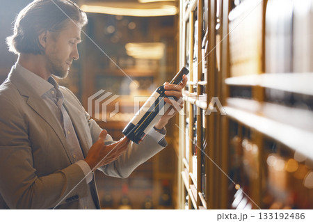 Experienced wine waiter holding wine bottle being in cellar on alcoholic drinks bottle shelf background. Sommelier takes notes scrutinizing various wine varieties. 133192486